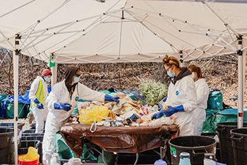 Workers in Personal Protective Gear at Table Under Canopy Sifting Through Trash from Opened Bags