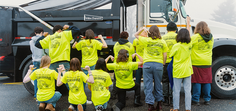 Group of Public Schools Students Wearing Yellow Shirts Pointing to the Logo on Back Standing Near Snowplow Truck